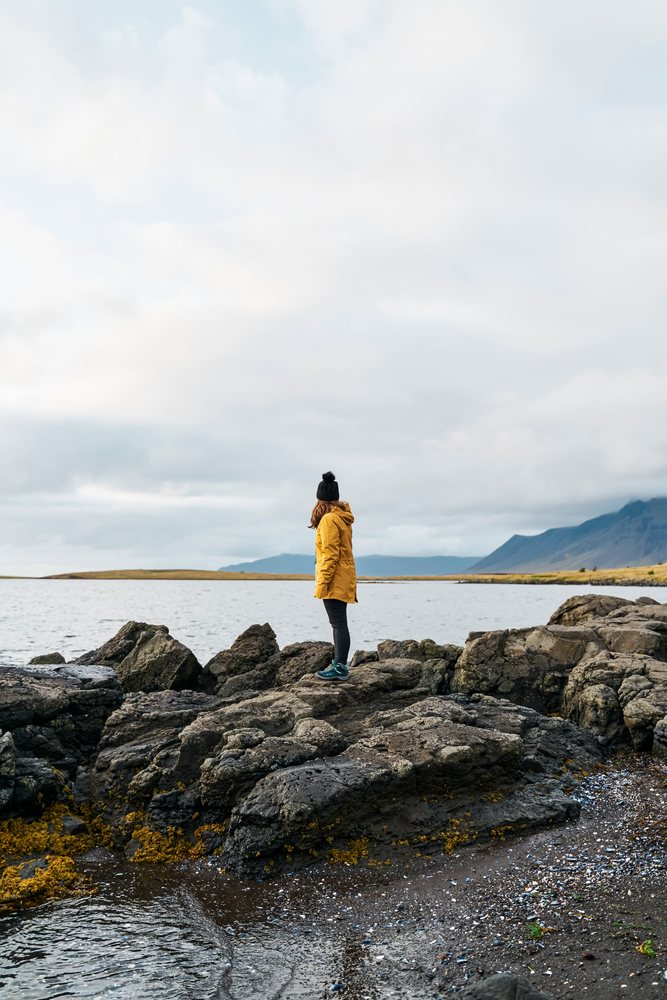 A white person stands on a rocky shoreline overlooking water
