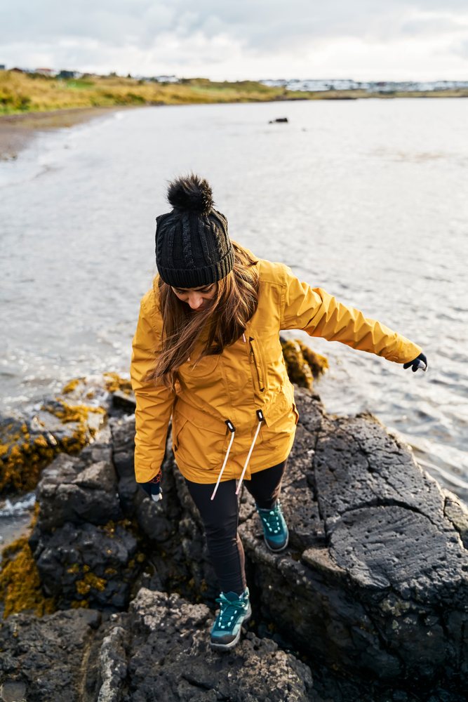 A white woman walks carefully on rocky shore wearing warm clothing