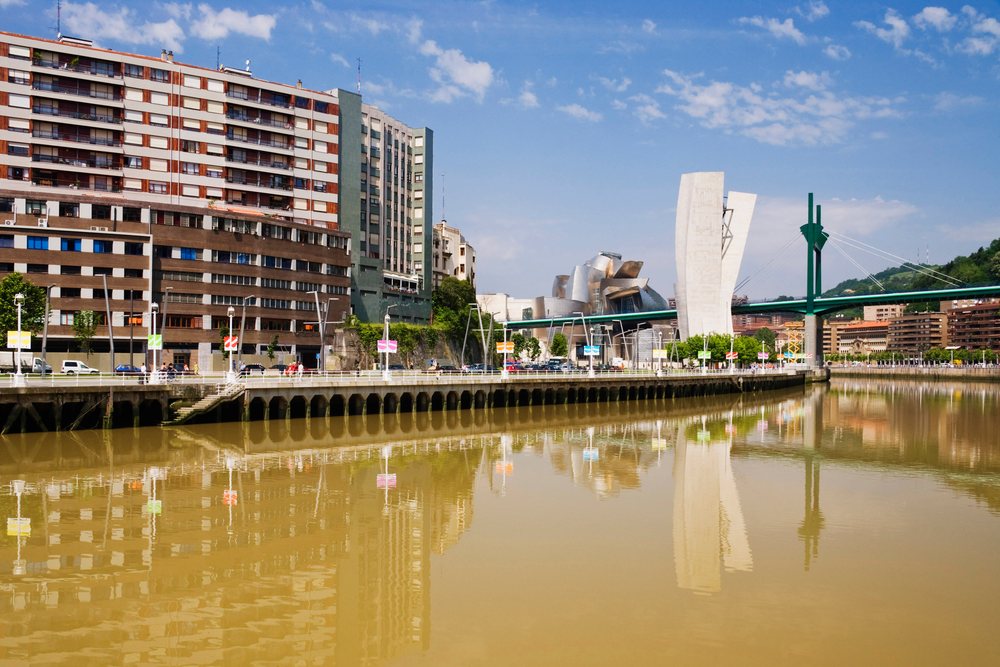 View of Nervion River with Guggenheim Museum, green bridge, and urban buildings reflecting in water on a clear day, Bilbao, Spain.