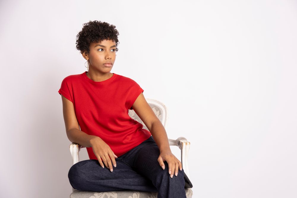 Woman with curly hair wearing a red shirt and dark jeans sits on a chair in a white studio setting, fashion, clothes, young culture.