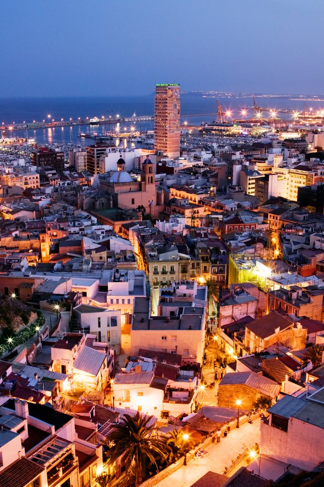 Night cityscape of Costa Blanca coastal city showing illuminated harbor, buildings, and hotel at dusk, Spain.