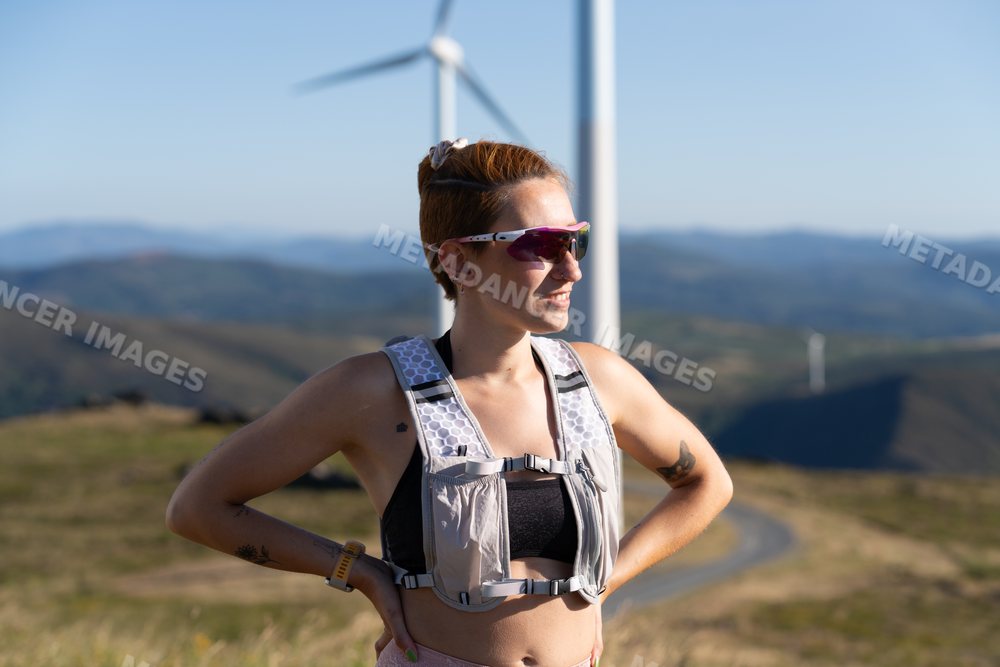 A white woman stands outdoors near wind turbines in the mountains