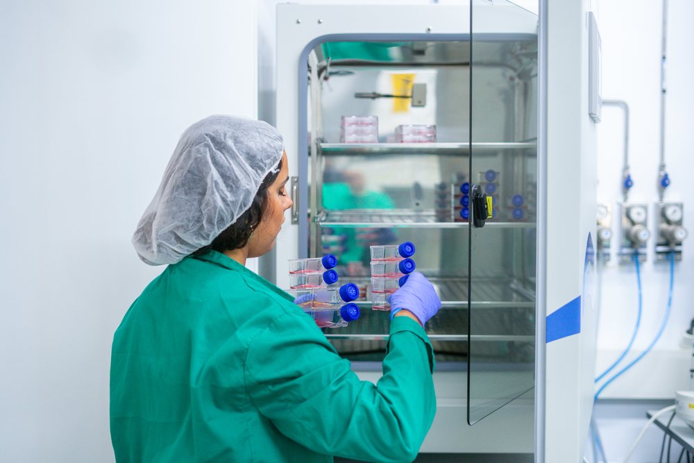 Laboratory technician wearing a green lab coat and blue gloves places biological samples into an open incubator in a sterile laboratory, research, biology, Seville, Andalucía.