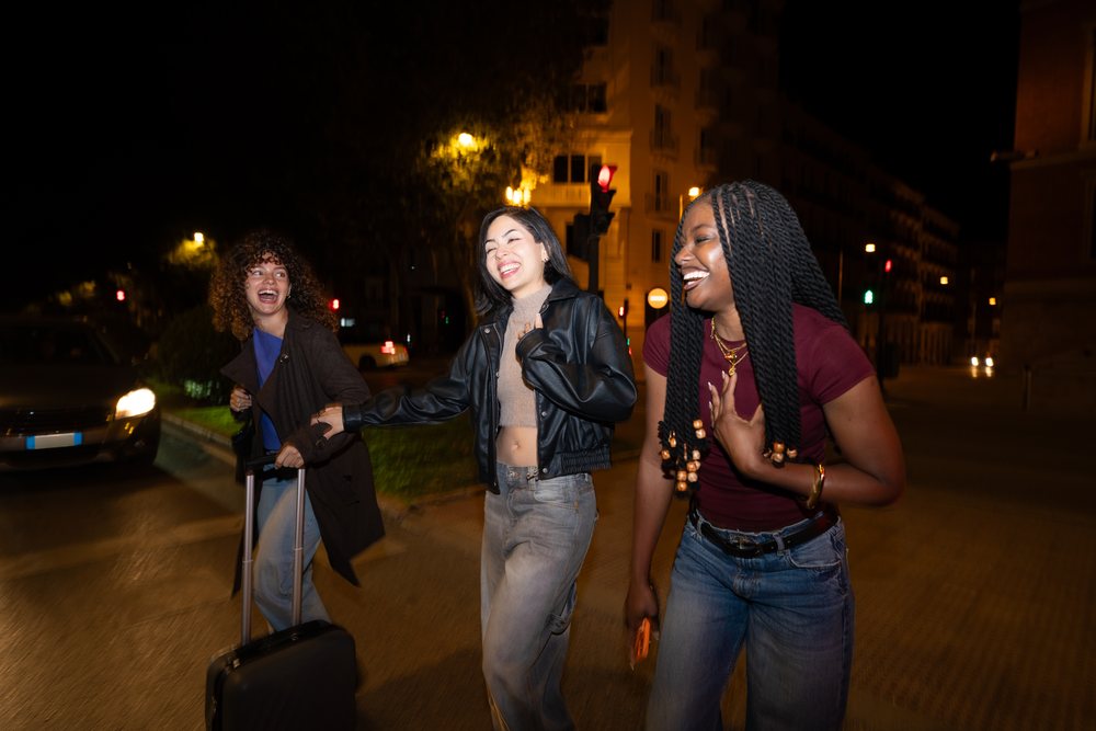 Three young women laugh and walk with a wheeled suitcase on an urban street at night, Madrid, Spain.