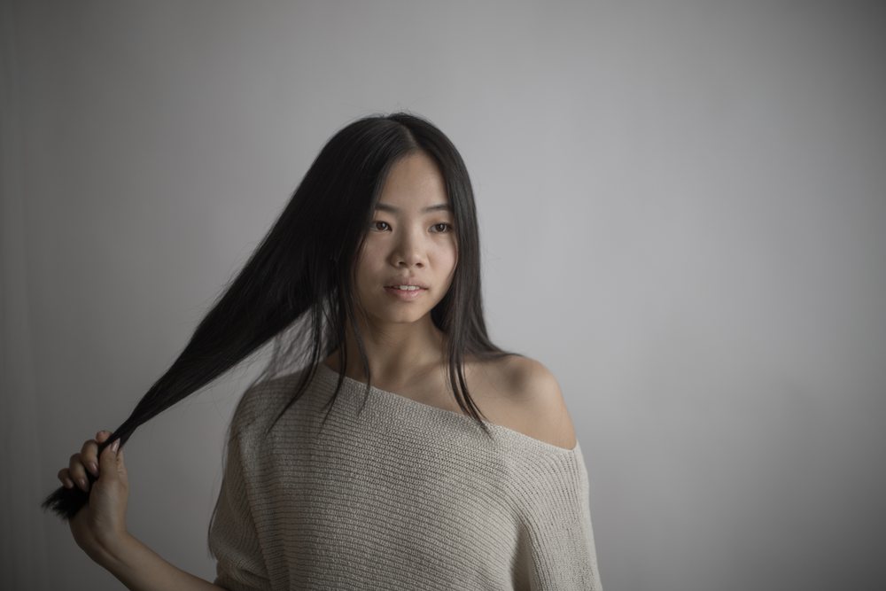 Portrait of a young Asian woman holding her long black hair wearing a gray off shoulder sweater against a neutral indoor background, Art, emotional.