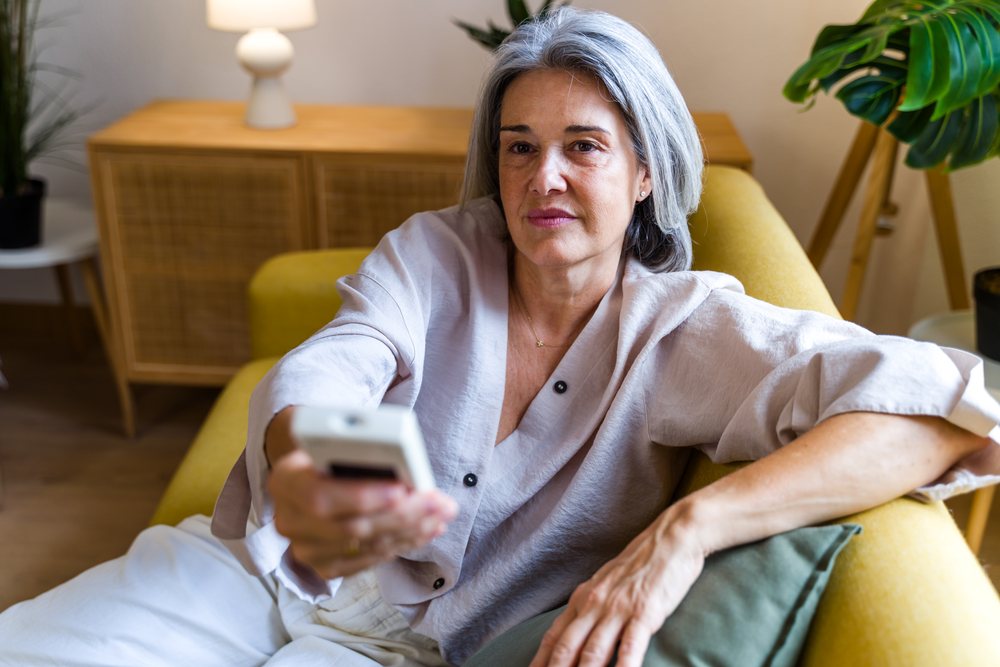Mature woman holding a remote control while sitting on a yellow couch in a contemporary living room with a houseplant and lamp nearby, Spain.