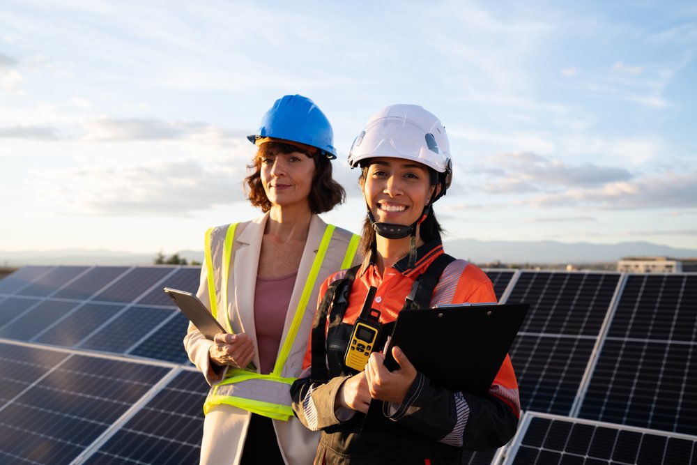 Two women engineers wearing helmets and safety gear stand by solar panels outdoors at sunset, Madrid, Spain.