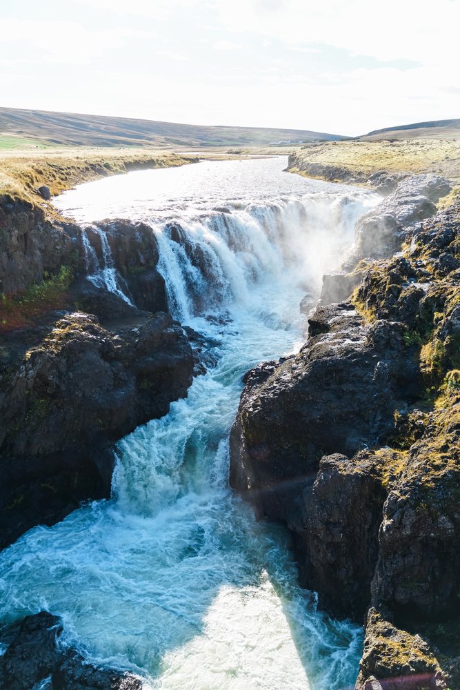 A waterfall cascades over rocky cliffs into a river surrounded by open land, Iceland.