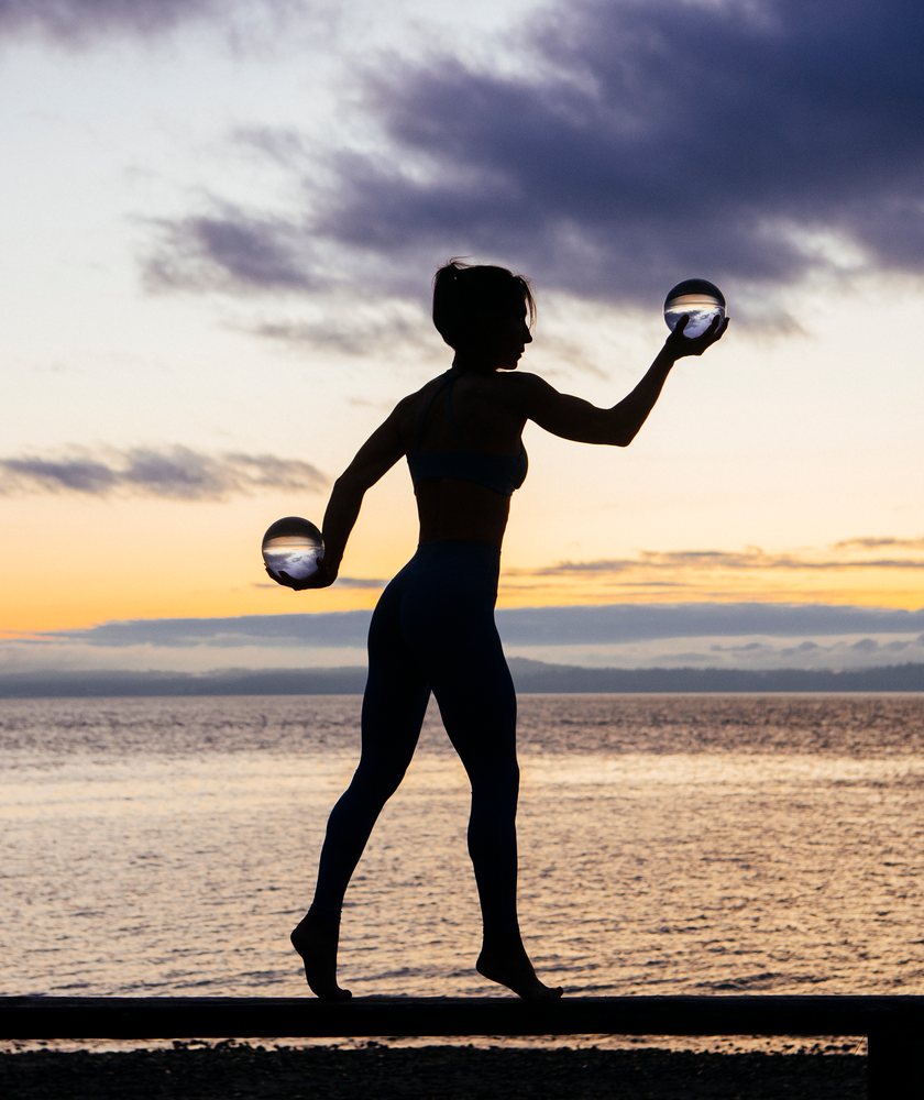 Silhouette of a woman holding two glass balls while balancing on a beam at sunset on a beach, WA, USA.