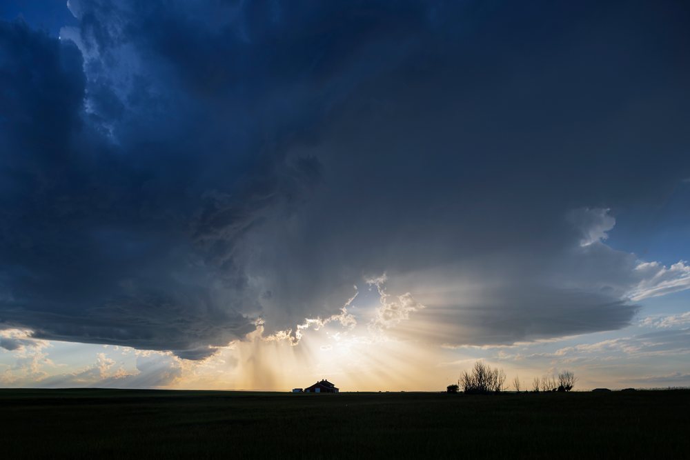 Silhouetted farm house under dark storm clouds with sun rays at Sunset over a rural field, Storm Chasing, Weather, Environment, High River, Alberta, Canada.
