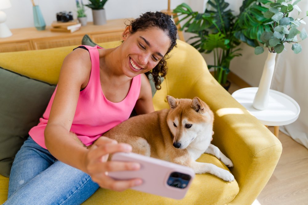 A woman in casual clothing takes a selfie with her medium-sized dog on a yellow sofa inside a cozy living room, Spain.