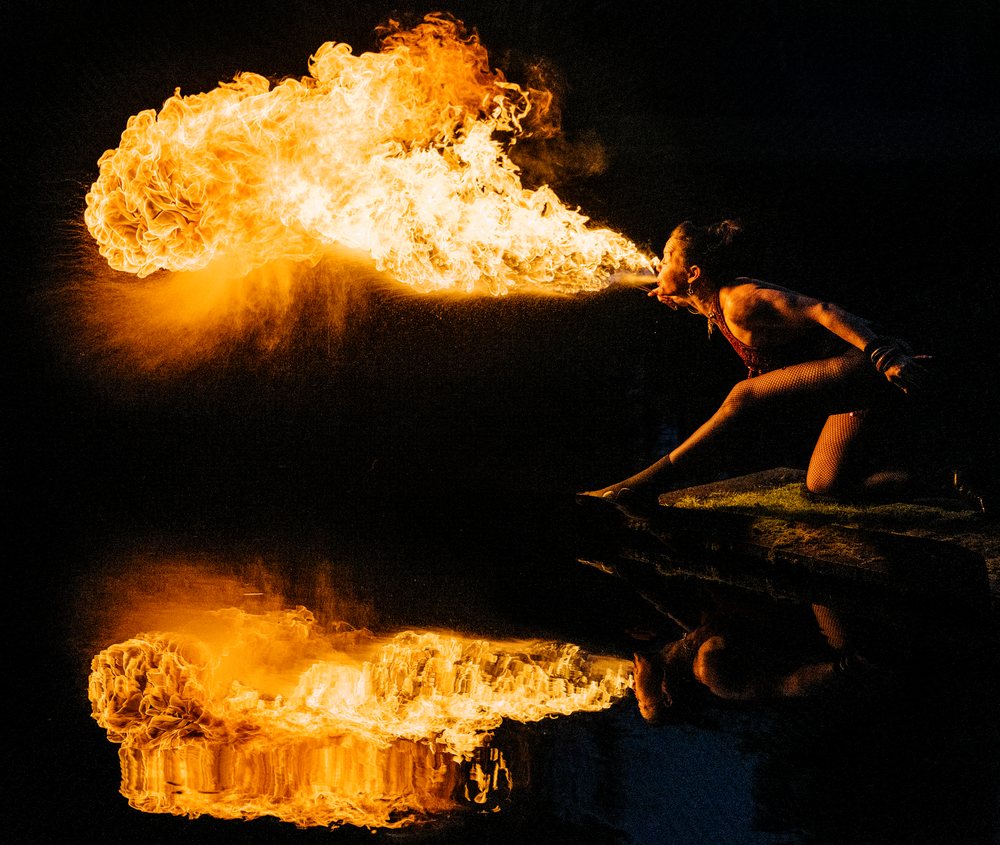 Woman fire performer breathes a large flame with dramatic reflection over water at night outdoors, artistry, artist, USA.