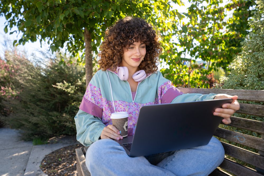 Woman with curly hair sitting on a wooden park bench outdoors, using a laptop and holding a coffee cup, Spain.