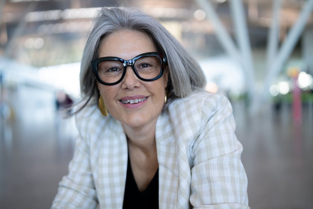 Middle-aged woman with gray hair, black glasses, and gold hoop earrings smiling indoors in a modern building, businesswoman, traveler, Spain, Madrid.