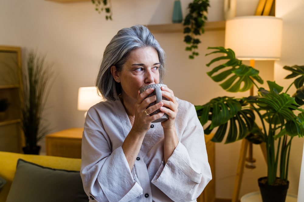Woman with gray hair drinks a hot beverage from a mug in a cozy, plant-filled living room, Spain.