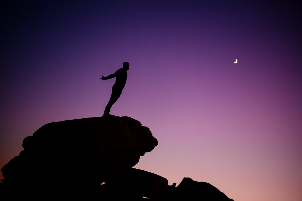 Silhouette of a man standing on a rock at twilight beneath a crescent moon, yoga, acrobatics, USA.