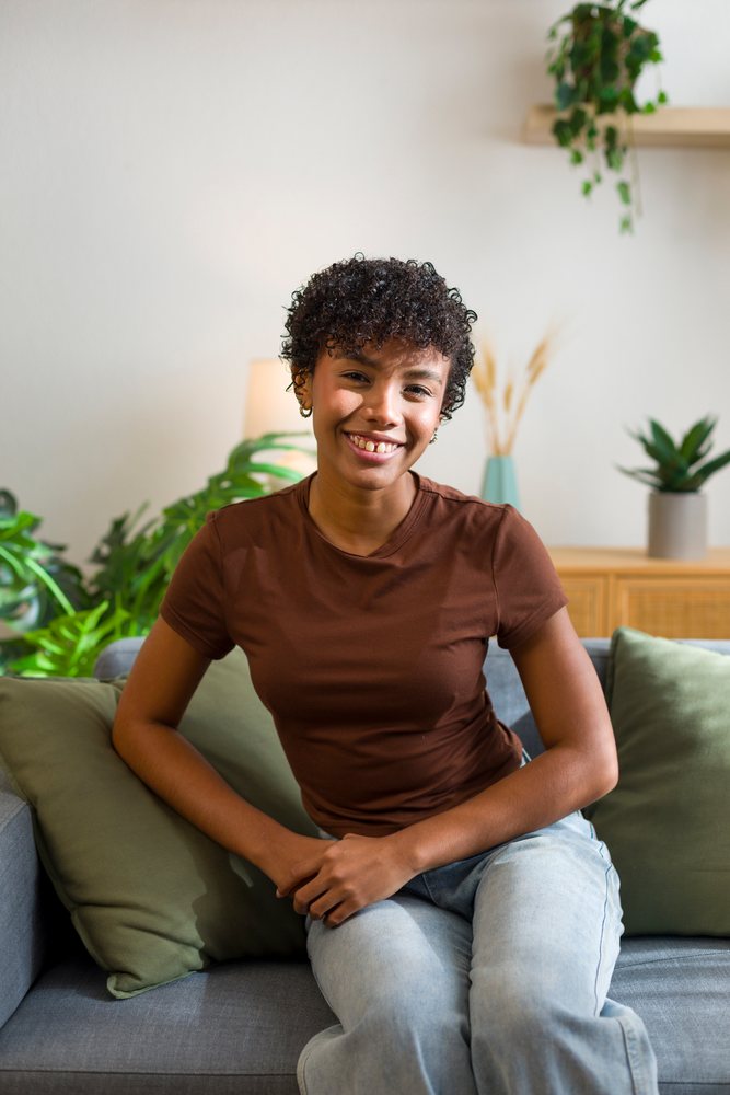 Young woman with curly hair smiles while sitting on a sofa with green cushions in a cozy living room with houseplants and wooden furniture.