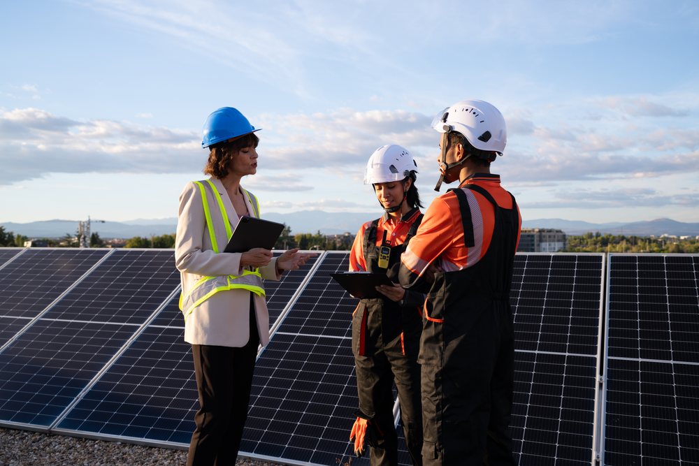 Three engineers in hard hats inspect solar panels during a consultation at a solar farm, with mountains in the background, Madrid, Spain.