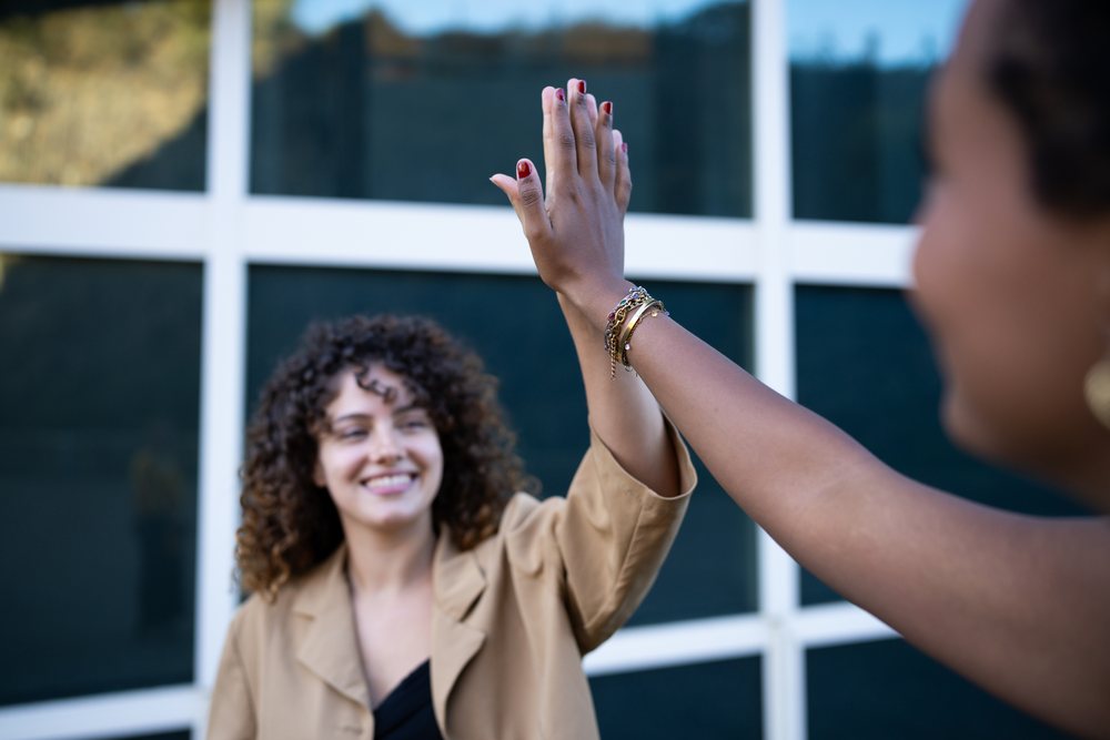 Two women outdoors near a glass wall share a high five, celebrating friendship and success, Spain.
