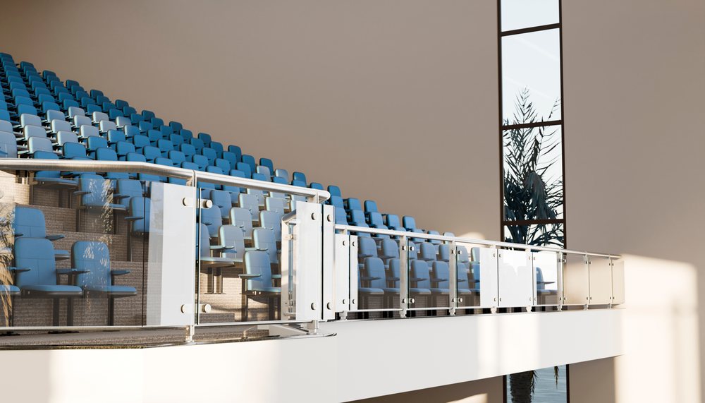Rows of blue cushioned seats with armrests face a glass panel railing inside a modern indoor stadium.