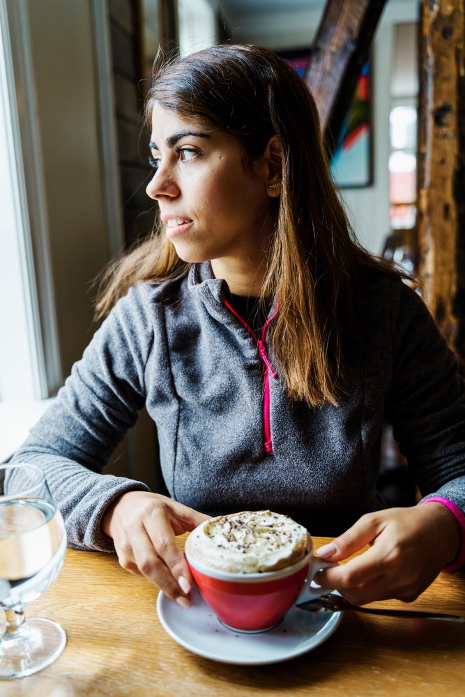 A woman wearing a gray fleece jacket holds a red cup of cappuccino topped with whipped cream and chocolate sprinkles while sitting indoors in a cafe, Iceland.