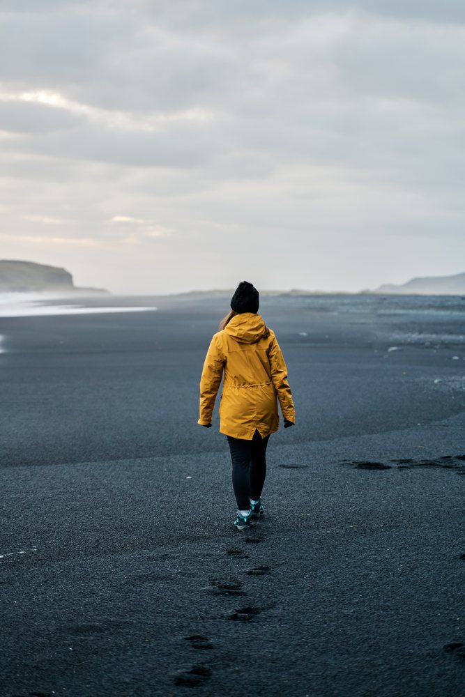 A white person walks on a black sand beach under an overcast sky