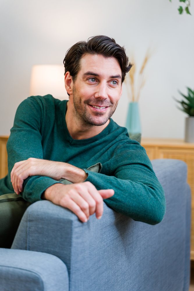 Man smiling while seated on a gray sofa indoors in a living room with plants and a lamp visible, Spain.