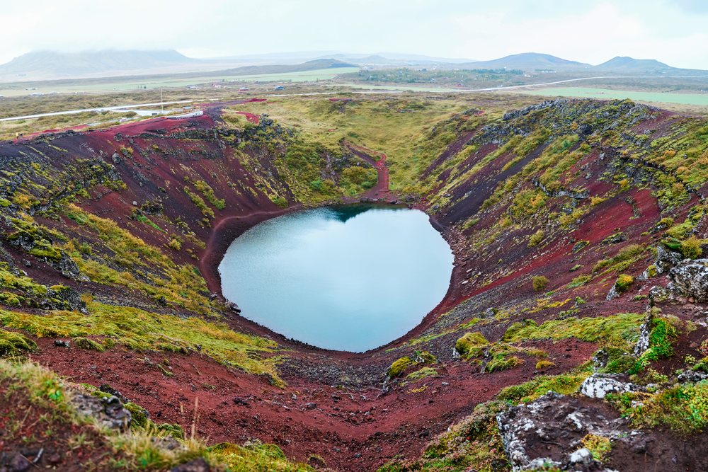 A scenic volcanic crater lake surrounded by red volcanic soil and green moss in a natural landscape, Iceland.
