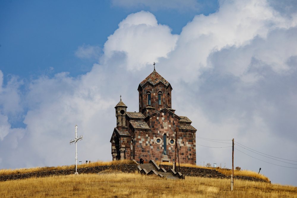 Stone St Sarkis Church with patterned masonry atop a dry grassy hill, Armenia under a partly cloudy sky, Tsakhunk.