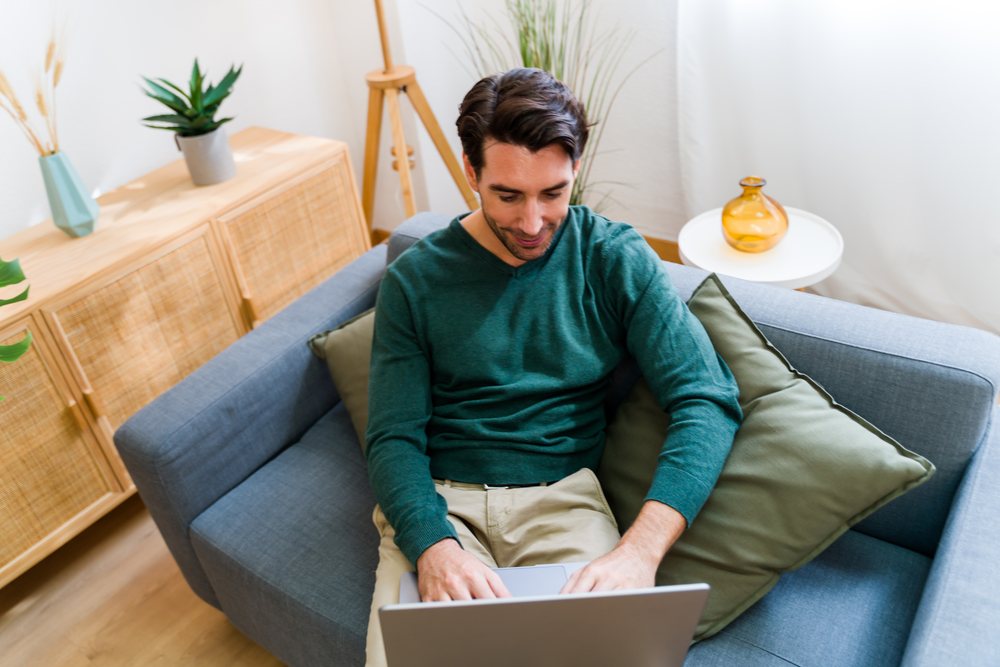 Man sitting on gray couch using a silver laptop in a bright living room with houseplants and wooden furniture, Spain.