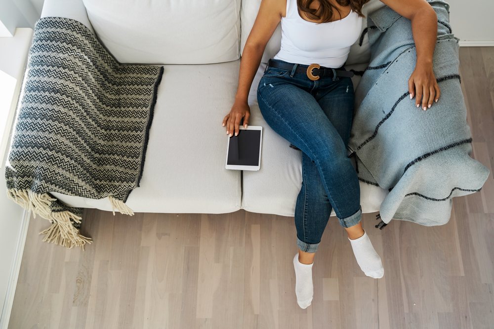 A white woman sits on a sofa holding a tablet indoors