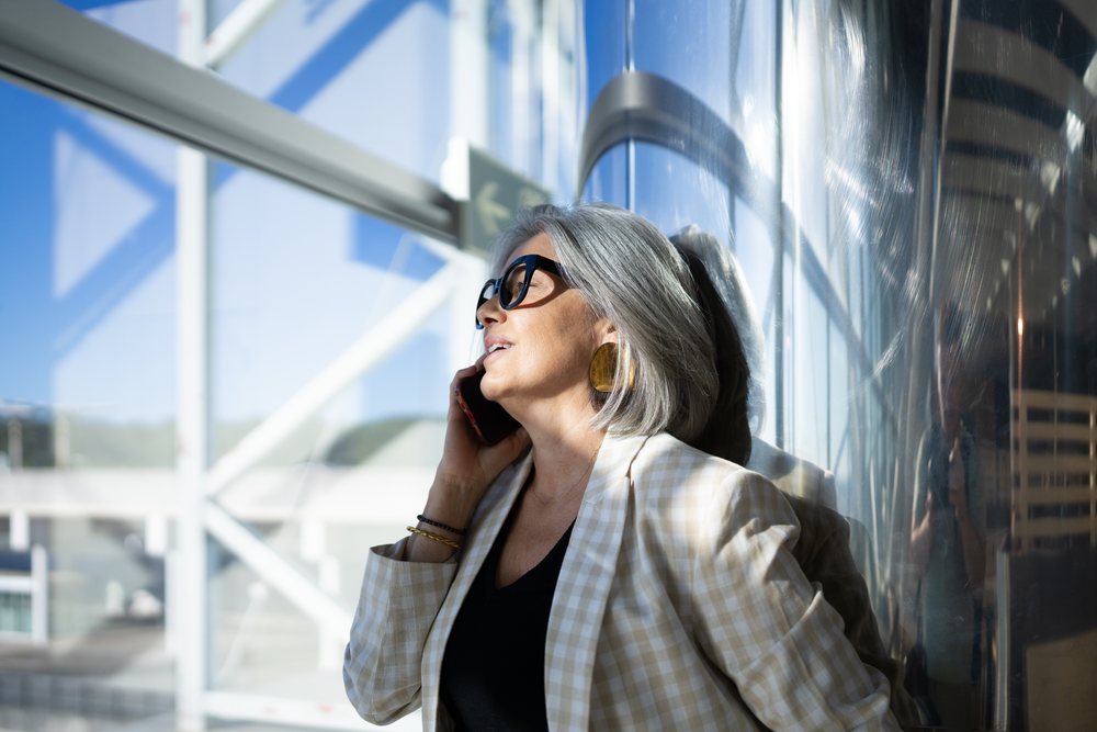Businesswoman with gray hair making a phone call indoors against a reflective metal surface in a modern building, Spain, Madrid.