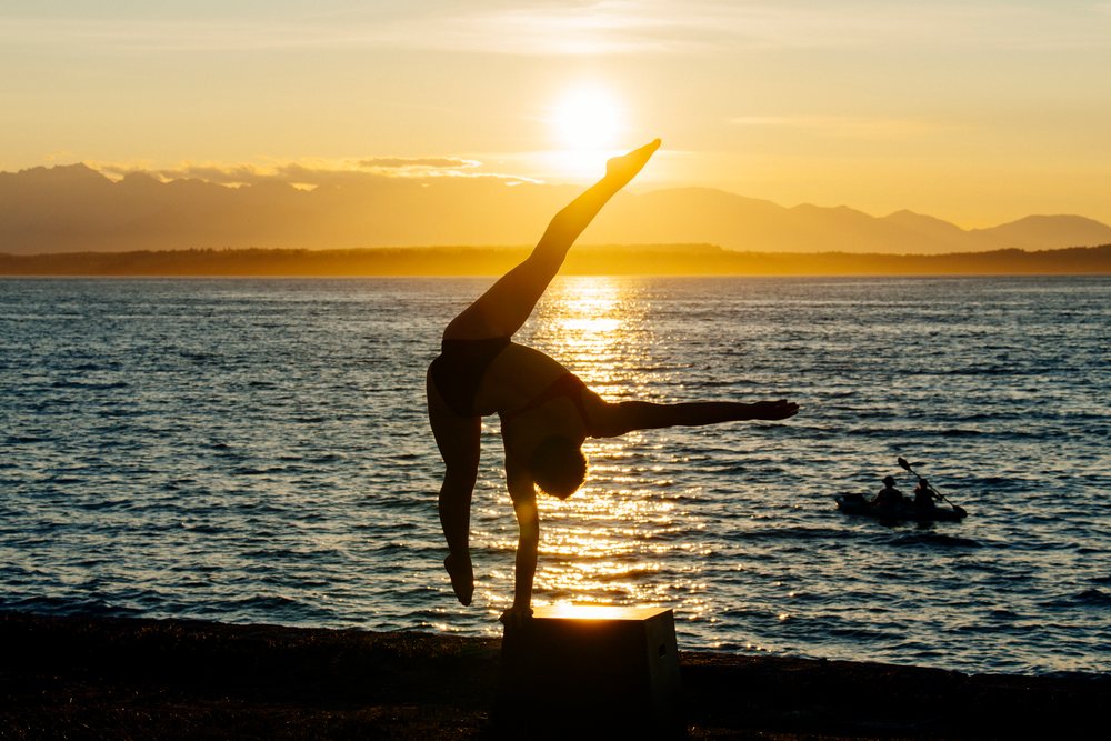 Silhouette of person balancing in handstand on a box by the ocean at sunset with kayaks on calm water, USA.