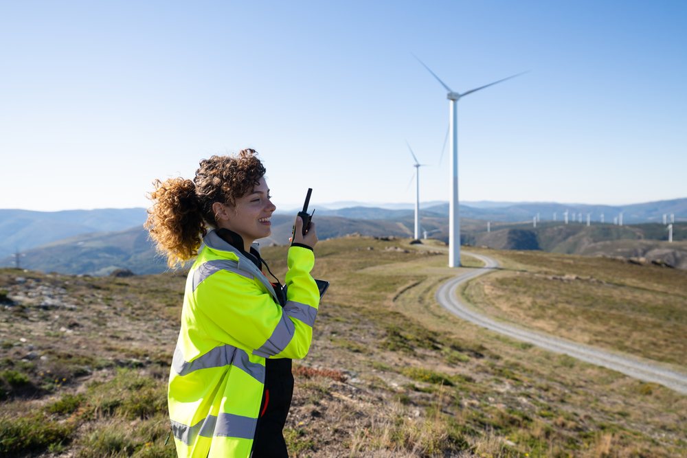 Smiling woman in a high visibility jacket uses a radio near wind turbines on a rural hillside, sustainability, renewable energy, Galicia, Spain.