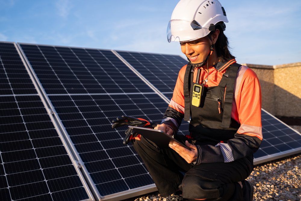 Woman solar energy technician wearing helmet and safety gloves, using a tablet to check solar panels outdoors at sunset, Madrid, Spain.