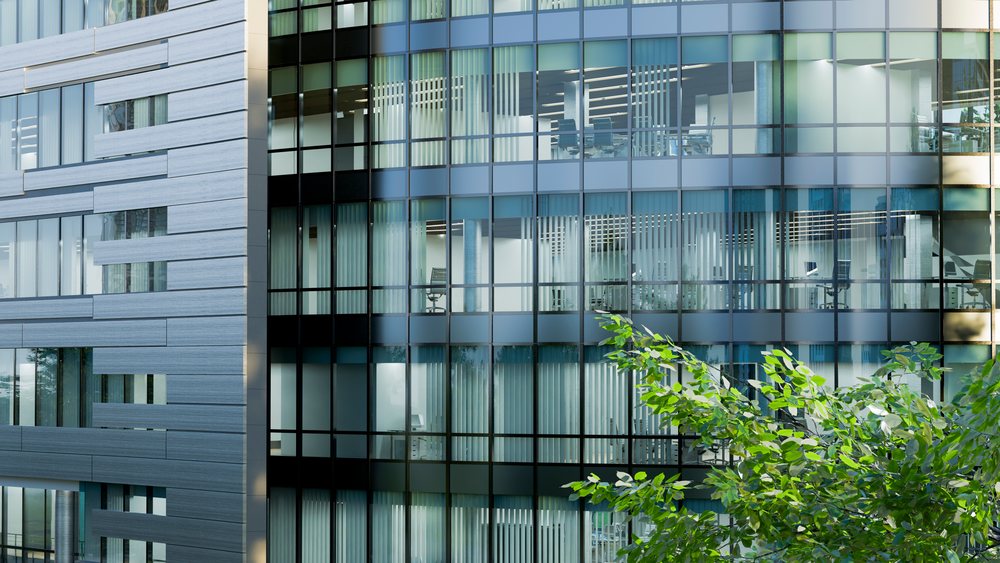 Modern office building with glass facade and visible conference rooms seen through trees in daylight.