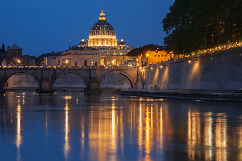 Illuminated St Peter's Basilica and an arched stone bridge over the Tiber River at dusk, Rome, Lazio, Italy.