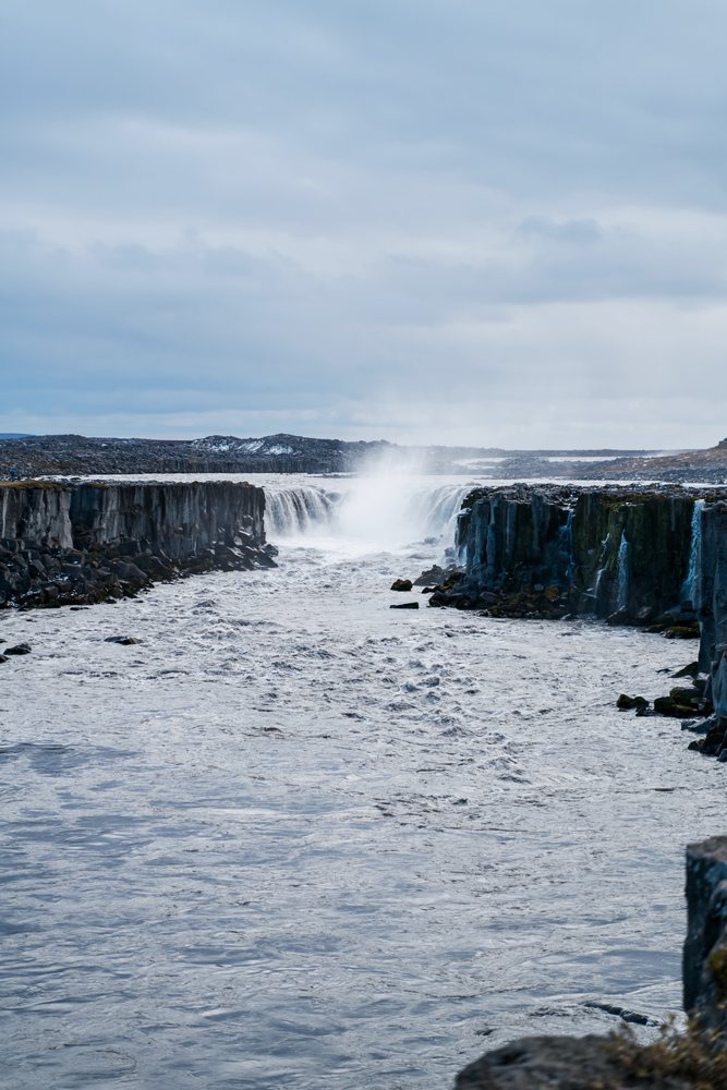 A powerful waterfall flows through basalt rock cliffs in a natural landscape in Iceland under an overcast sky.