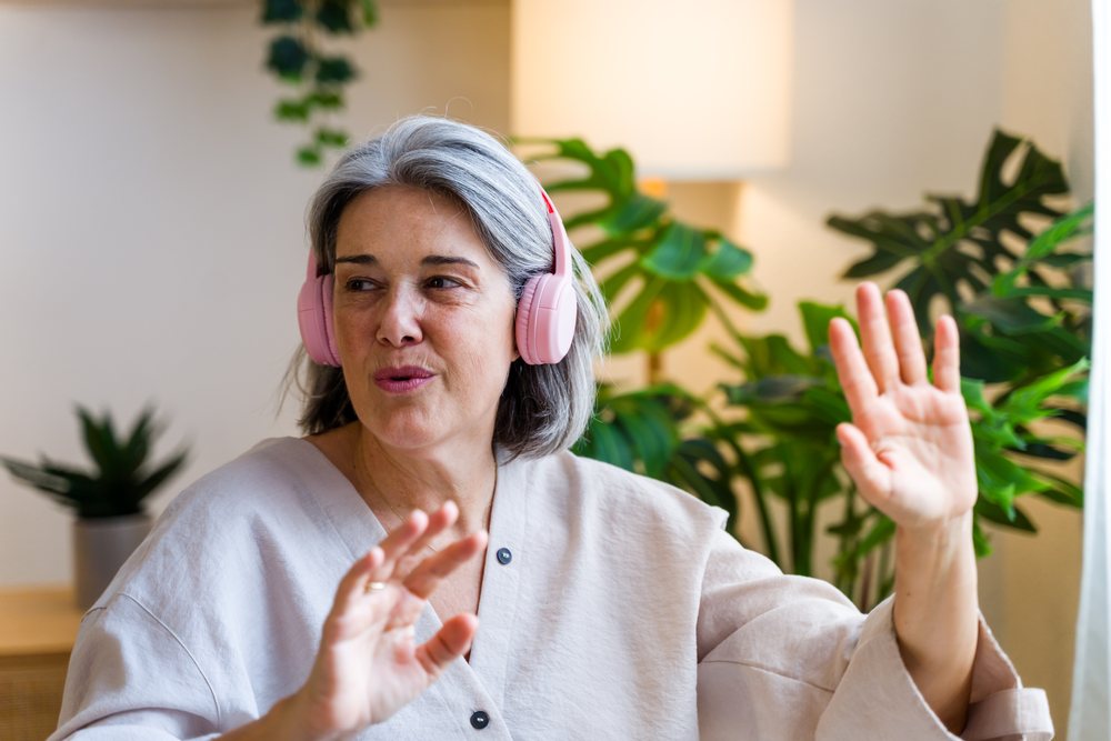 Mature woman with gray hair wearing pink headphones gestures while enjoying music indoors near houseplants and a lamp, Spain.