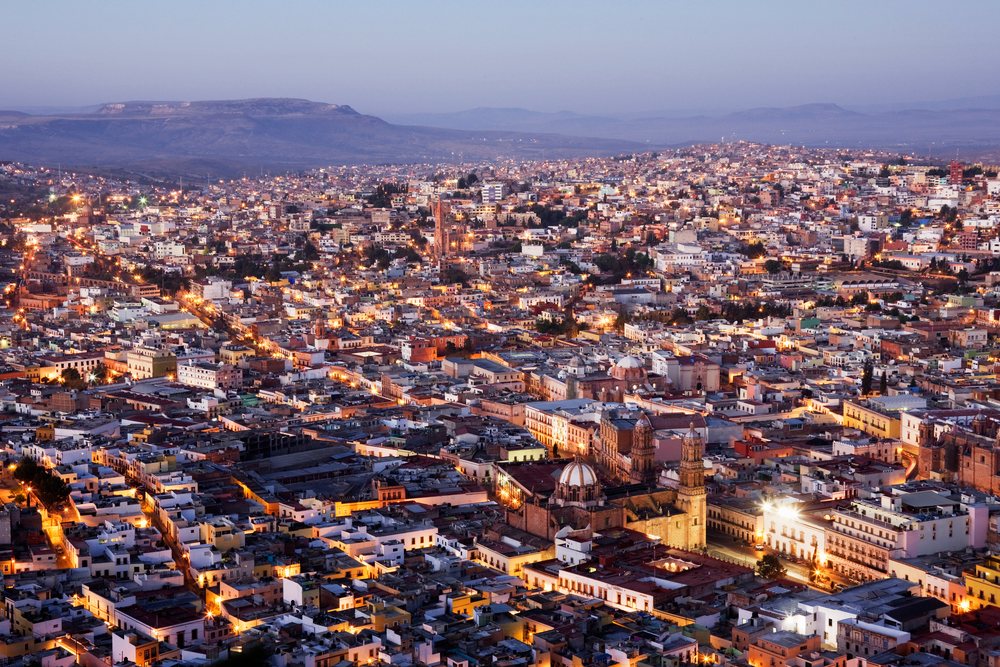 Cityscape of Zacatecas at dusk showing illuminated historic buildings and mountain backdrop, Mexico.