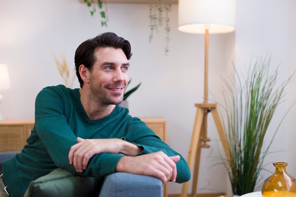 Man smiling and sitting on a couch in a cozy living room with a floor lamp and houseplants, technology, young adult, home.