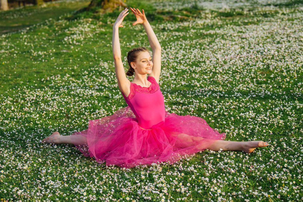 Ballerina in pink tutu and leotard performs a split on a sunny grassy field dotted with daisies, USA.
