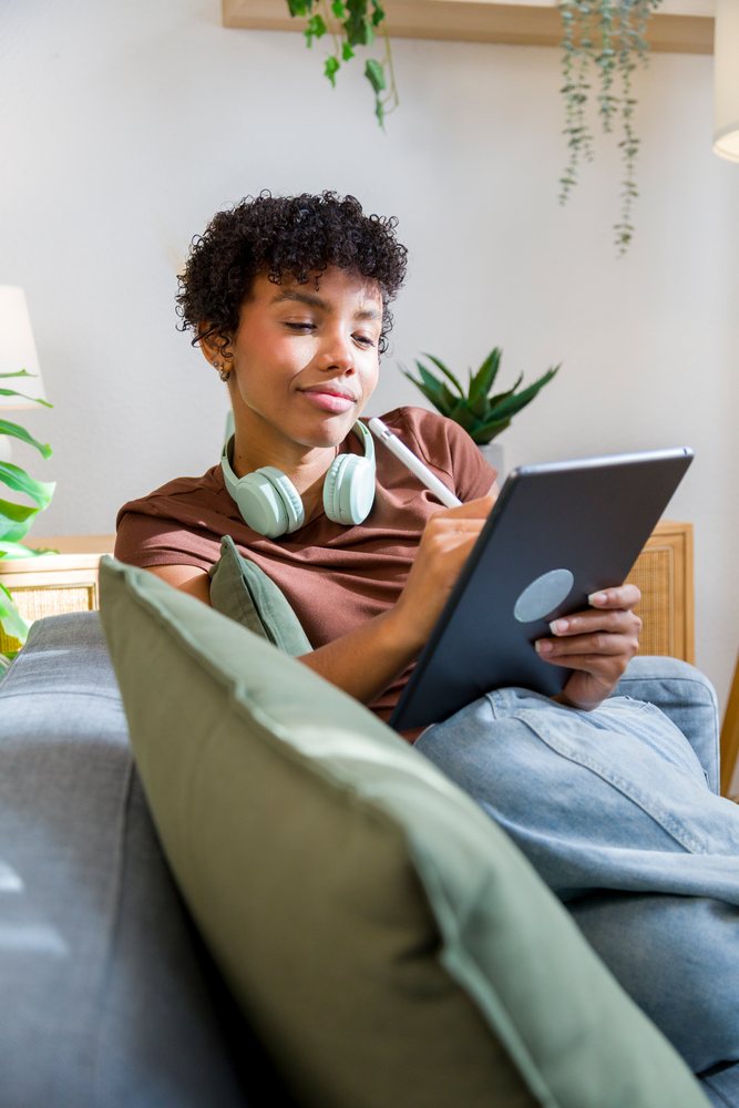 Young woman using a tablet with a stylus while sitting on a sofa indoors with plants nearby.