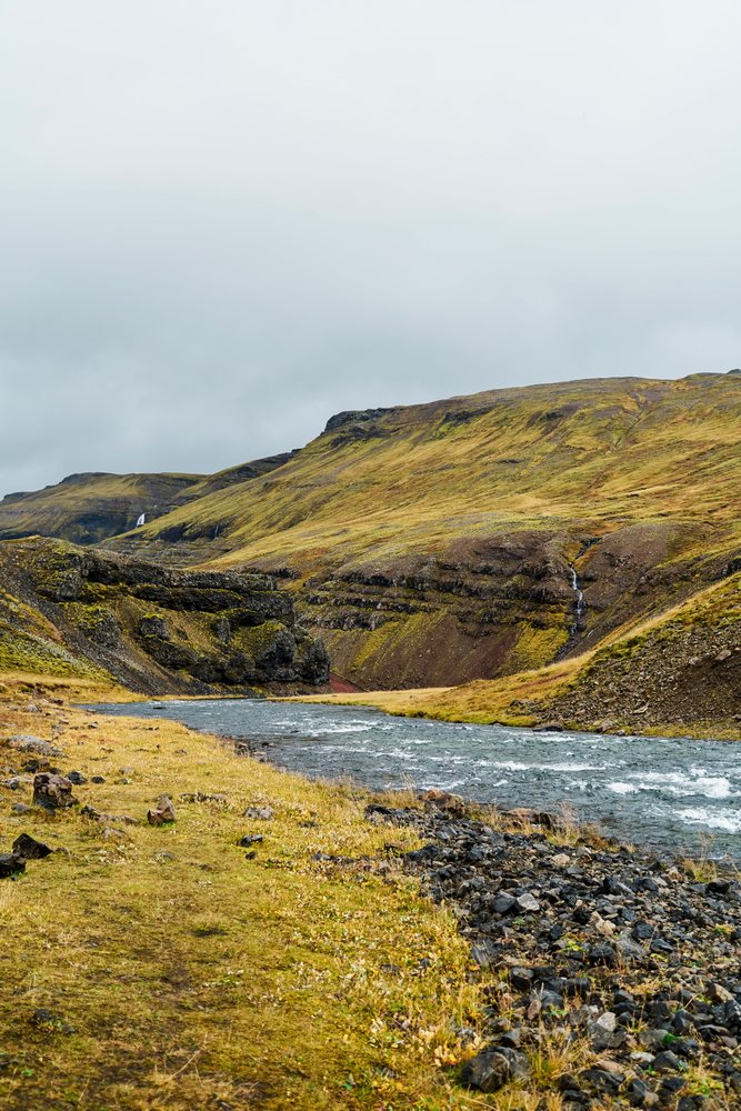 A river flows through a rocky valley with hills and waterfalls under an overcast sky, Iceland.
