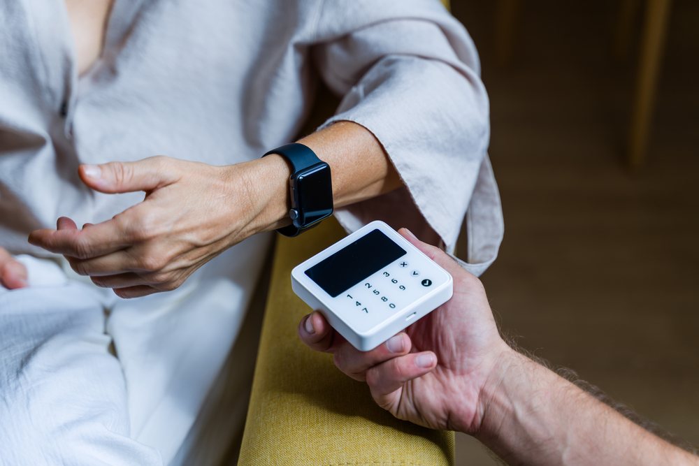 Person wearing a smartwatch on the wrist holding a small digital device with a numeric keypad indoors, Spain.