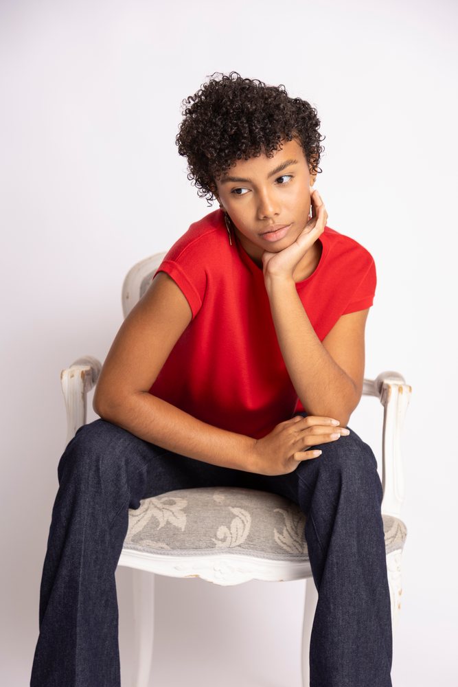Thoughtful woman with curly hair in red shirt and blue jeans sits on an decorative chair in a studio setting, fashion, clothes, young adult culture.