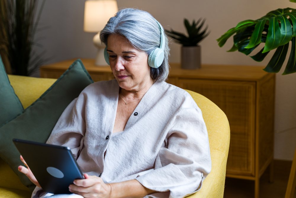 Gray-haired woman wearing headphones uses a tablet while seated on a yellow chair with green cushions in a living room, Spain.