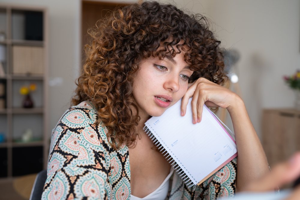 Woman with curly hair holding a notebook thoughtfully in a home interior setting, Spain.