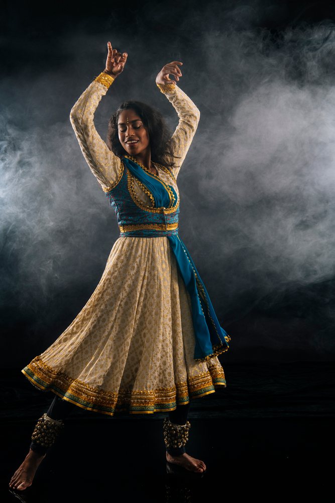 Woman performs classical Indian dance in a white dress with gold embroidery, blue sash, and ankle bells against a dark studio background with smoke, expressing joy and spirituality.