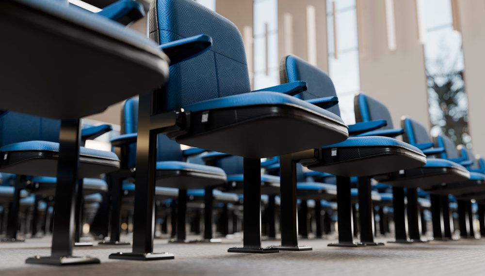 Multiple blue padded chairs with armrests and metal frames are arranged in rows inside a modern conference hall with large windows and hanging lights.